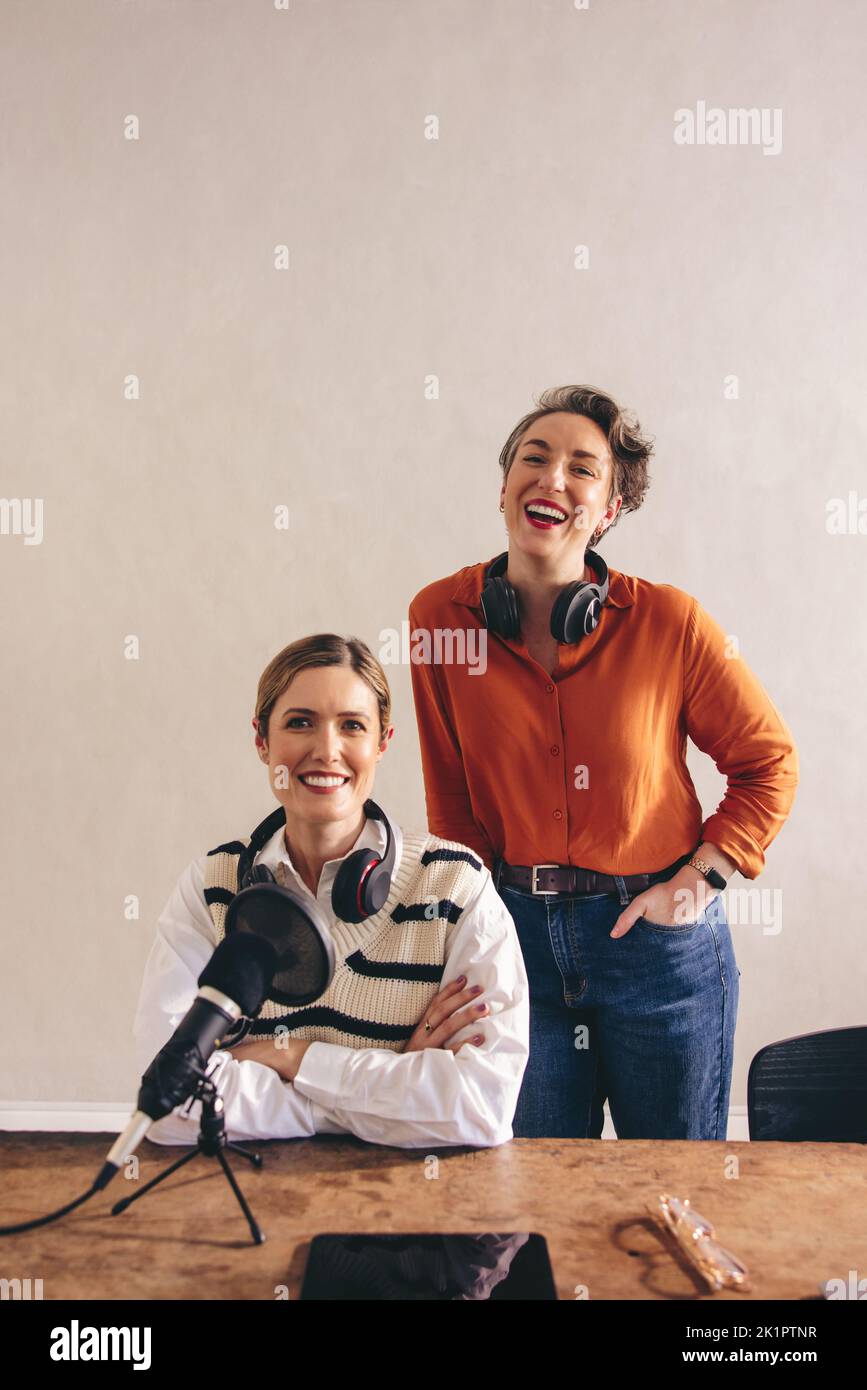 Deux baladoteurs femelles souriant à la caméra avec des casques autour de leur cou. Des femmes heureuses co-accueillant une émission audio dans un studio à domicile. Ambiance chaleureuse et agréable Banque D'Images