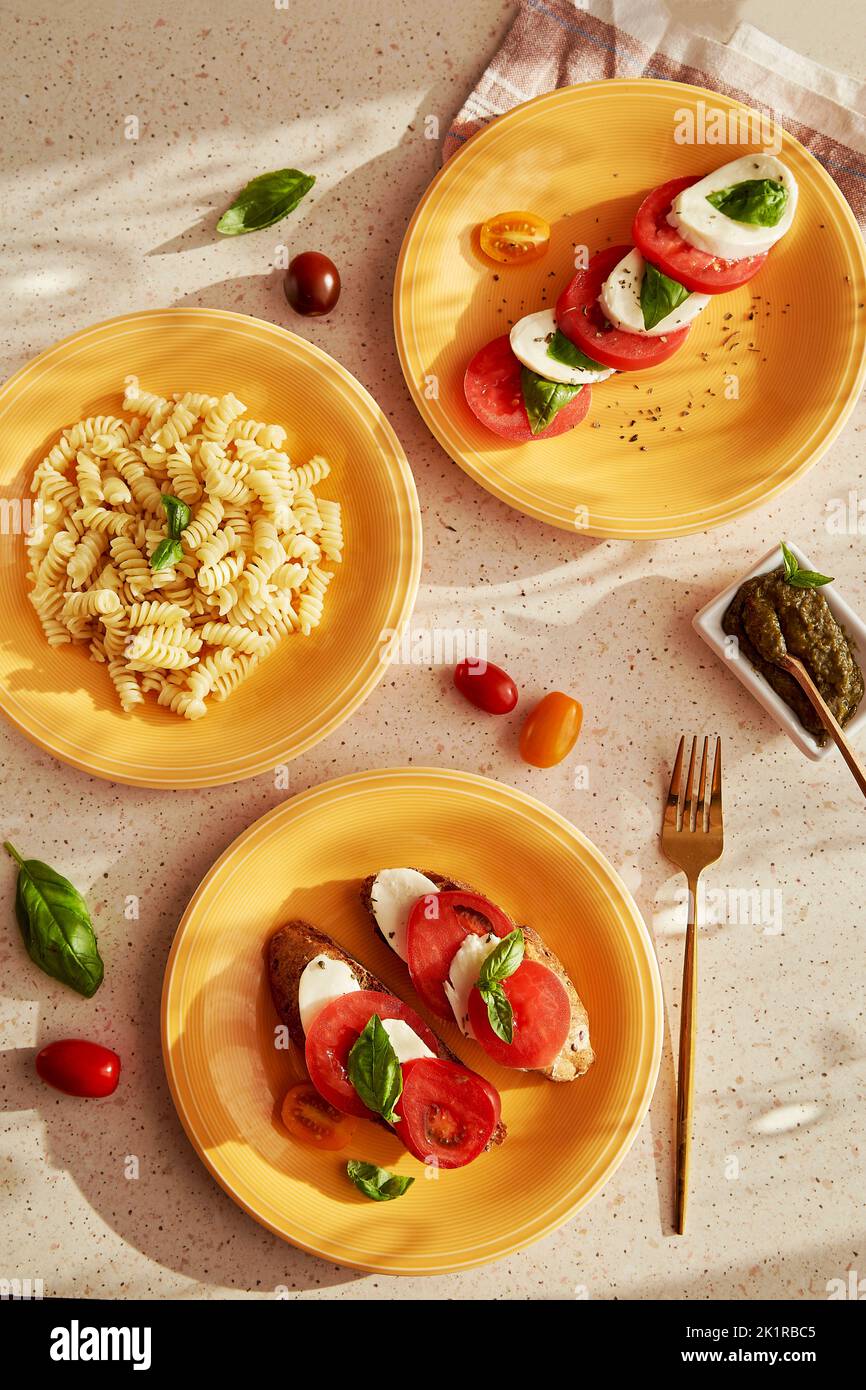 Assortiment de plats italiens avec pâtes fusilli, salade de caprese - tomates, mozzarella, basilic et pesto. Hors-d'œuvre au pain avec salade de caprese sur pla orange Banque D'Images