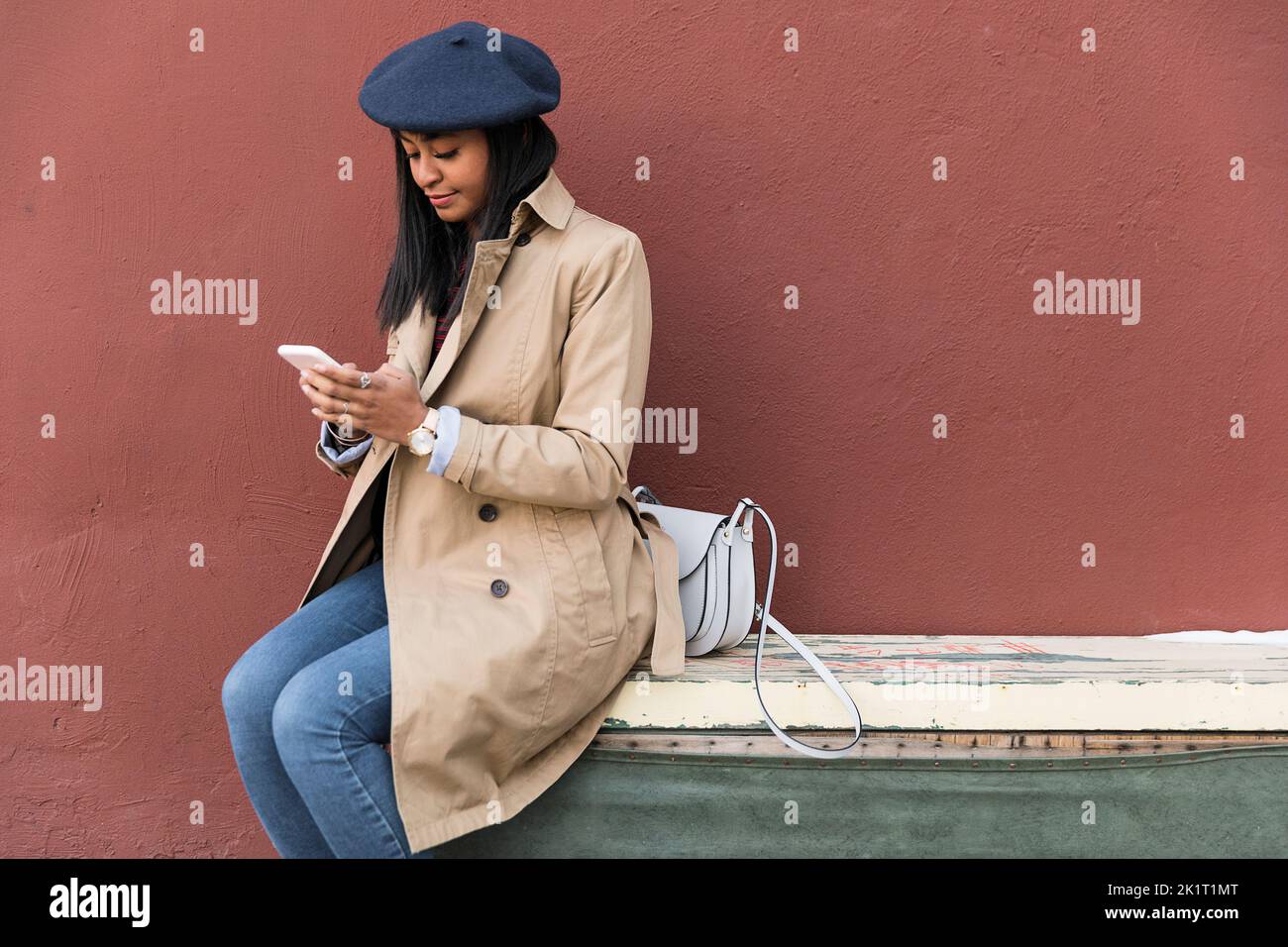 Jeune femme en béret et en trench coat textant avec un smartphone Banque D'Images