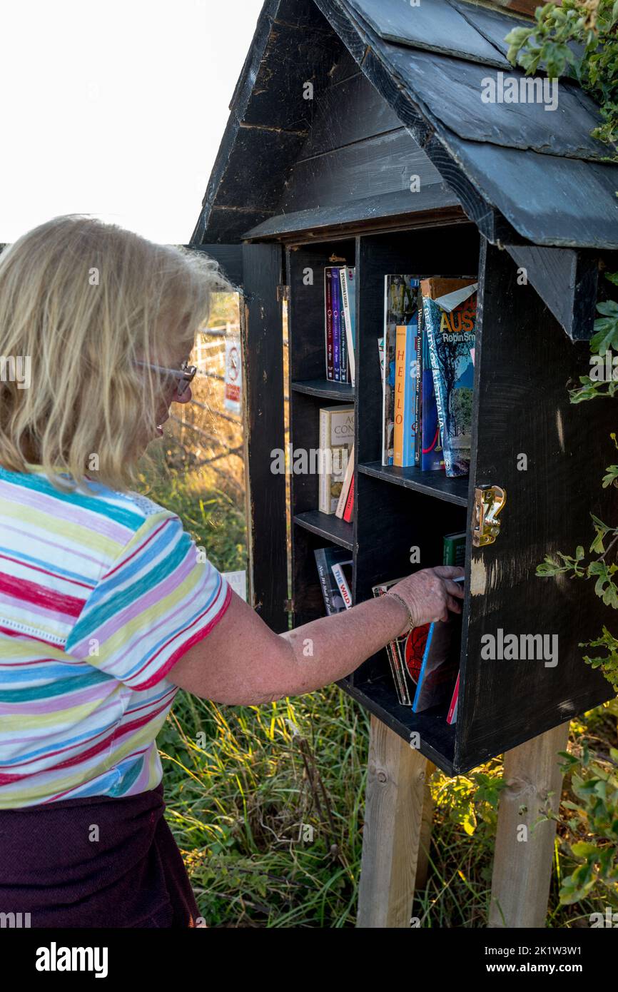 Femme prenant un livre d'un livre de la partie prendre un livre-laisser un livre d'un abri en bois peint noir dans le village côtier d'Embleton, Northumberland. Banque D'Images