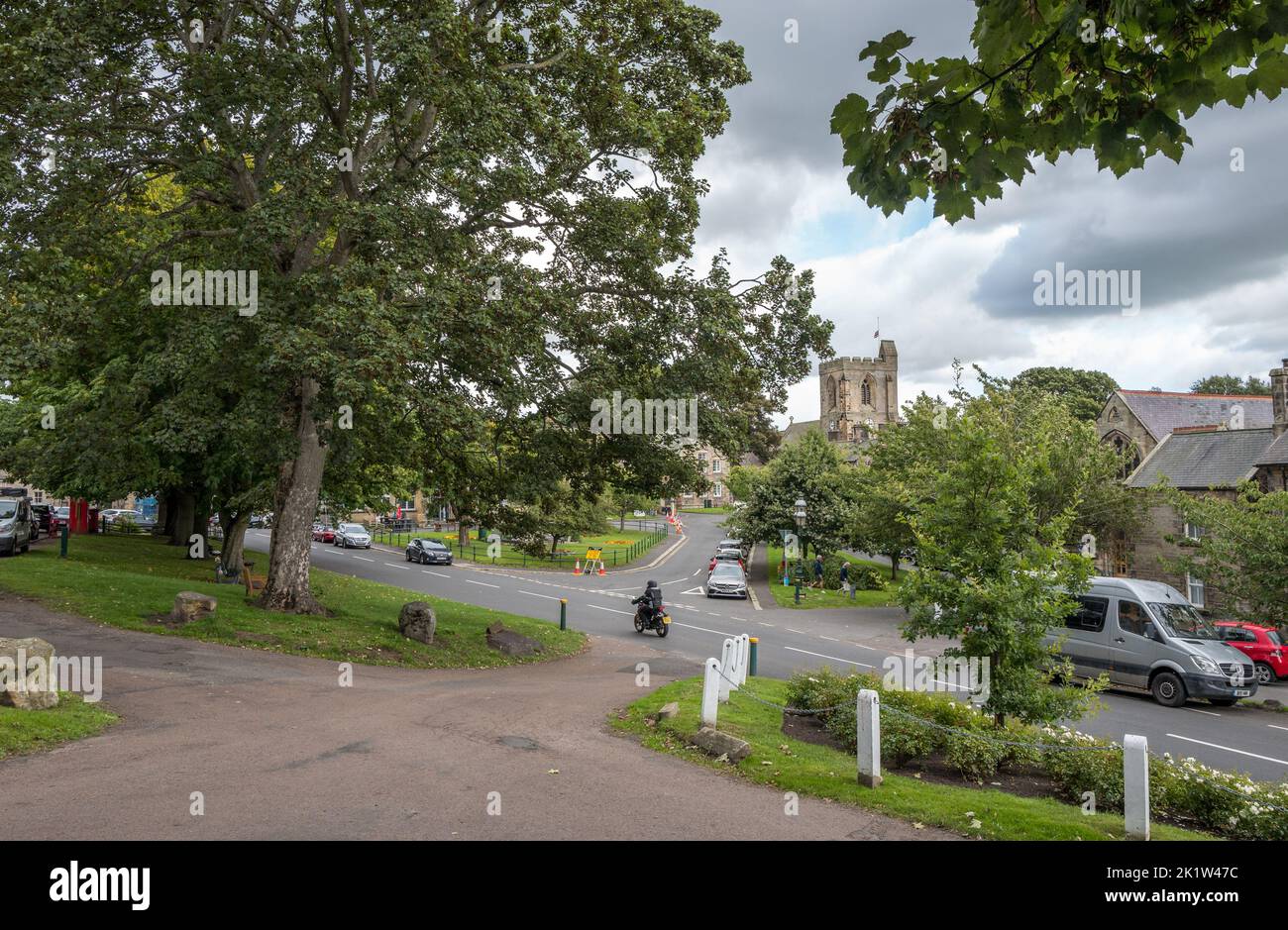 L'arbre borde la route principale à travers dans la petite ville marchande de Rothbury dans Northumberland, Angleterre avec l'église de tous les Saints en arrière-plan. Banque D'Images