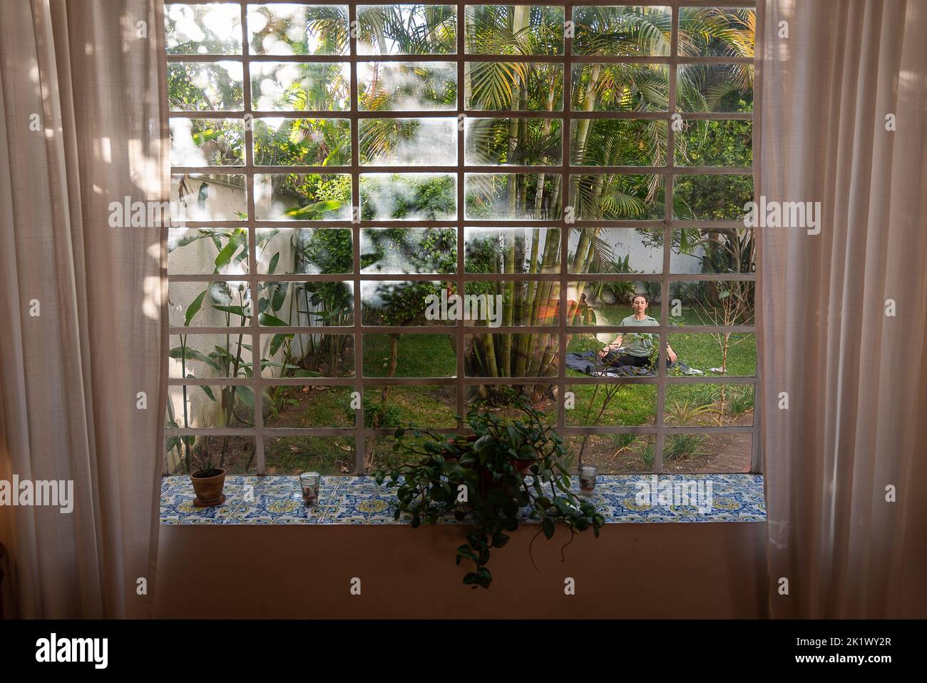 La femme pratique le yoga dans l'arrière-cour entouré de plantes et d'arbres, capturés par la fenêtre de la maison avec des vibes tropicales et des palmiers Banque D'Images