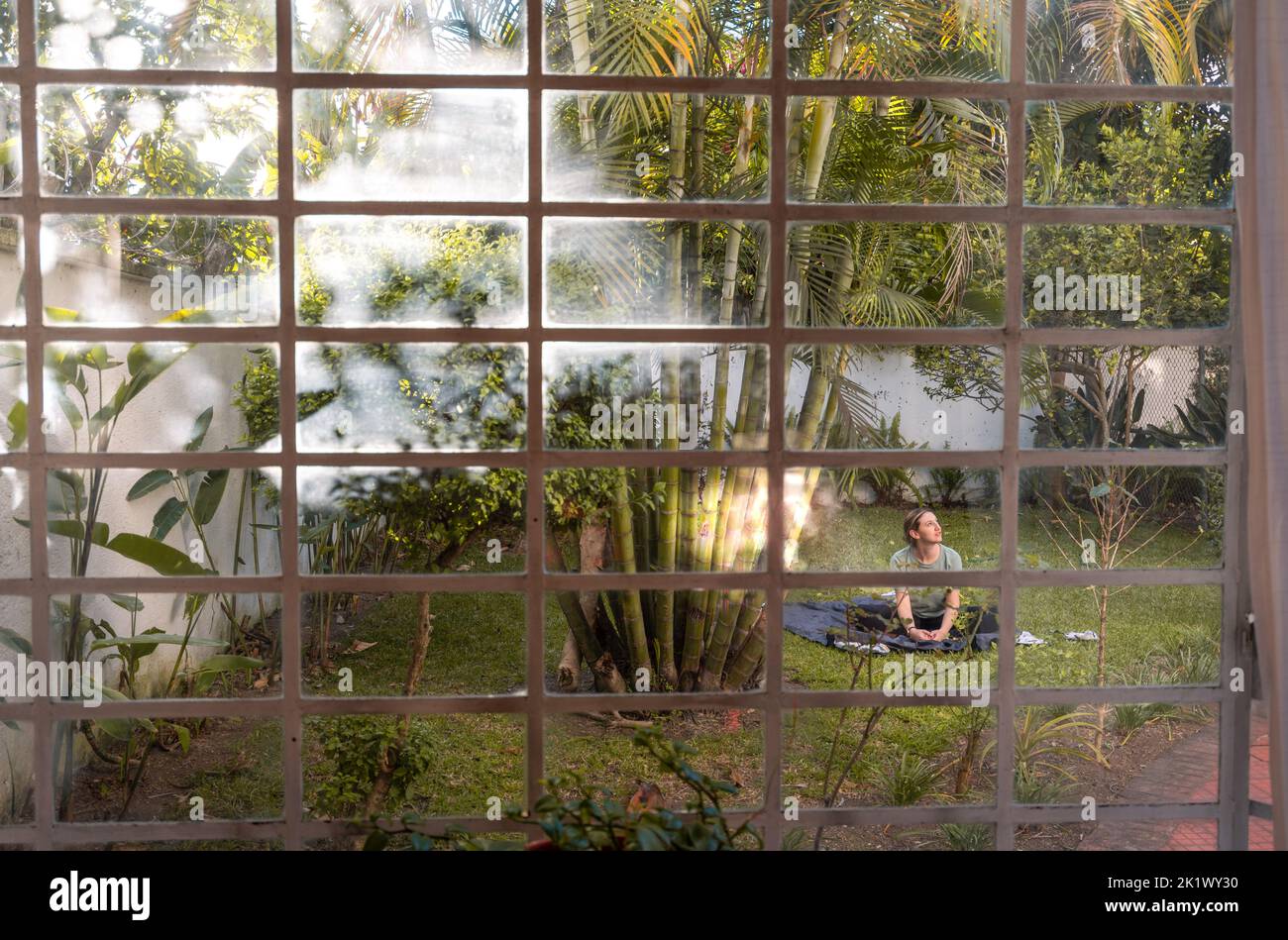 La femme pratique le yoga dans l'arrière-cour entouré de plantes et d'arbres, capturés par la fenêtre de la maison avec des vibes tropicales et des palmiers Banque D'Images