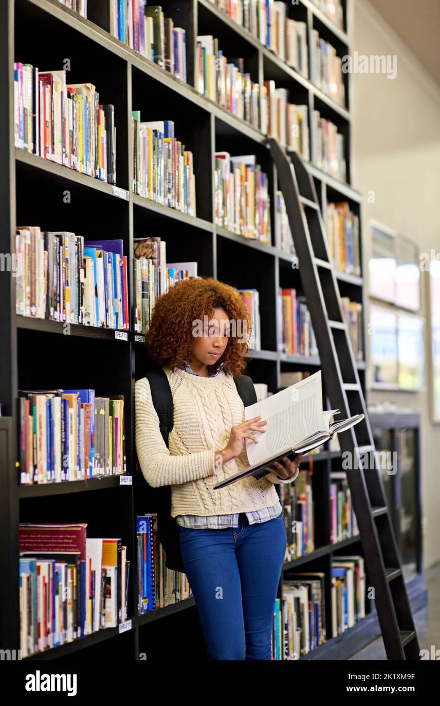 La connaissance est libre à la bibliothèque... il suffit d'apporter un conteneur. Une jeune femme étudie à partir d'un livre tout en se tenant près d'une bibliothèque. Banque D'Images