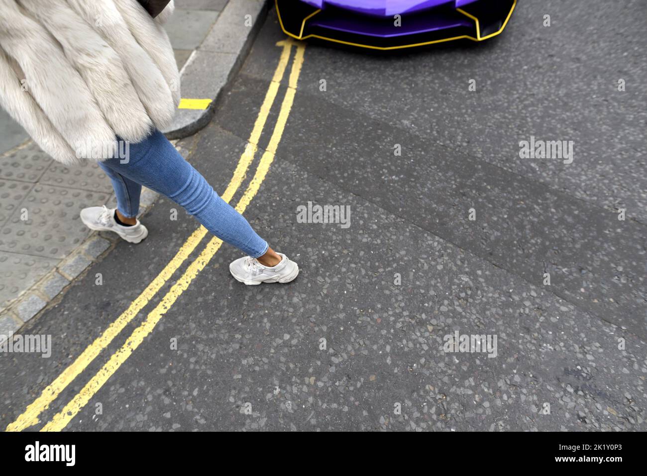 Londres, Angleterre, Royaume-Uni. Femme en Jean et un manteau de fourrure marchant devant une Lamborghini garée Banque D'Images