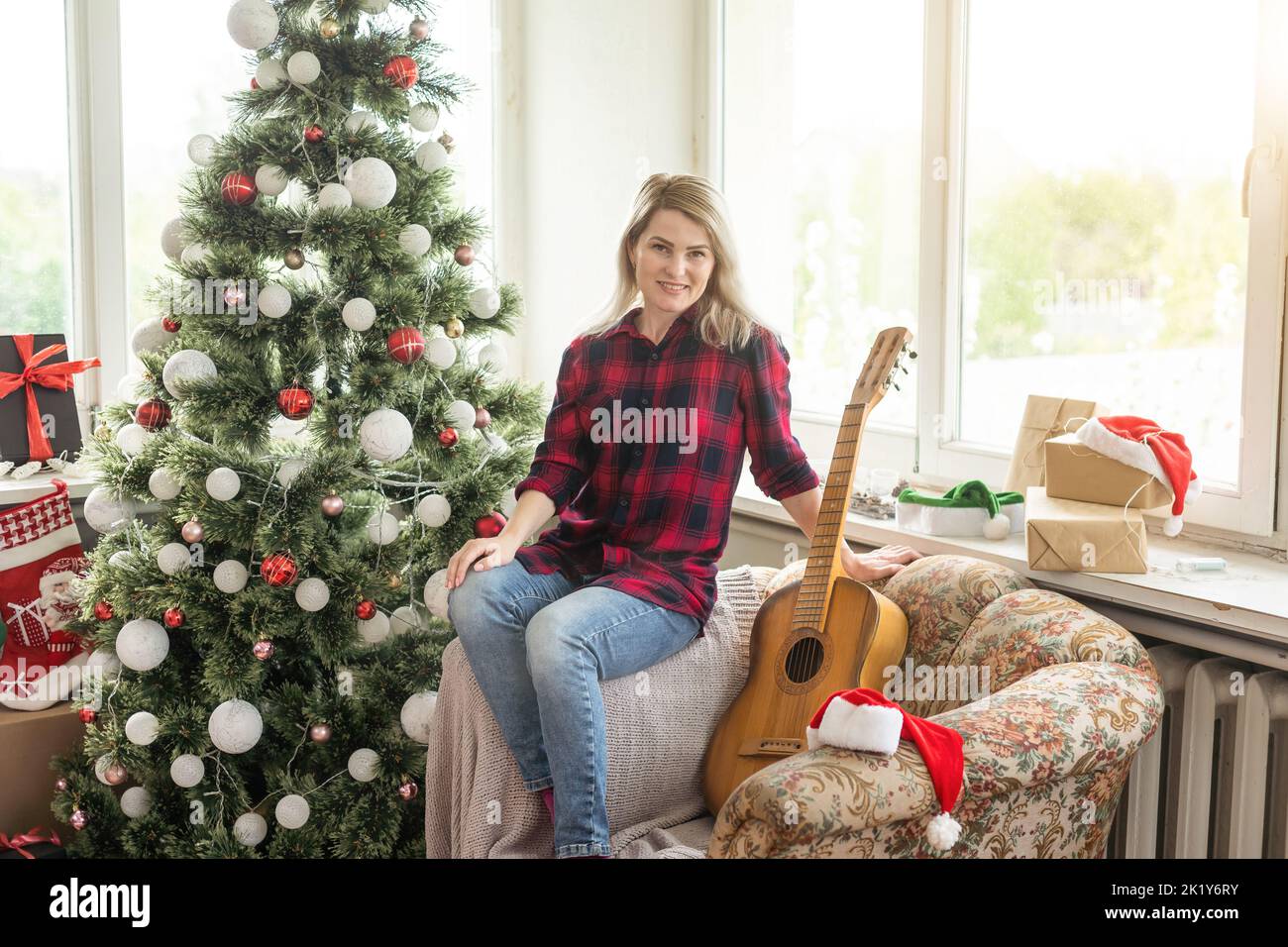 Belle fille se trouve près de l'arbre de Noël décoré de boules et de lumière et jouant la guitare et le chant. Jeune belle femme souriante jouer Banque D'Images