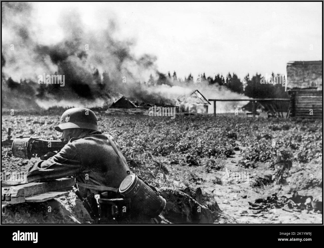 WW2 OPÉRATION BARBAROSSA action contre-partisane de Whermacht soldats allemands nazis sur le front oriental. Au premier plan, un soldat allemand en position de tranchée, tirant une mitrailleuse MG-34 avec un magasin de batterie. Un village en feu à l'arrière-plan. 1941 Banque D'Images