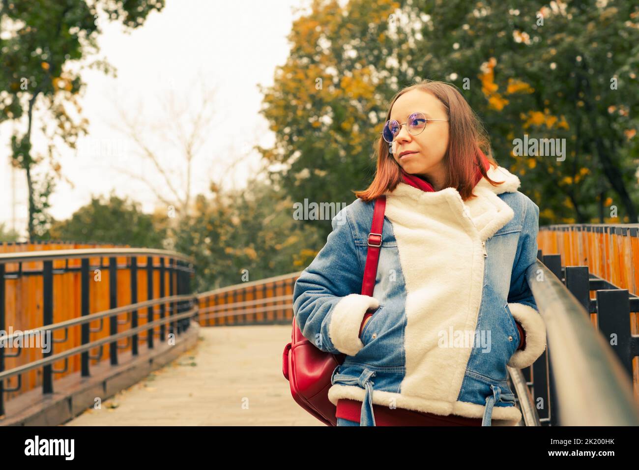 Jeune belle femme en lunettes à cheveux courts en veste denim chaude, en regardant loin, debout sur la passerelle par temps frais Banque D'Images