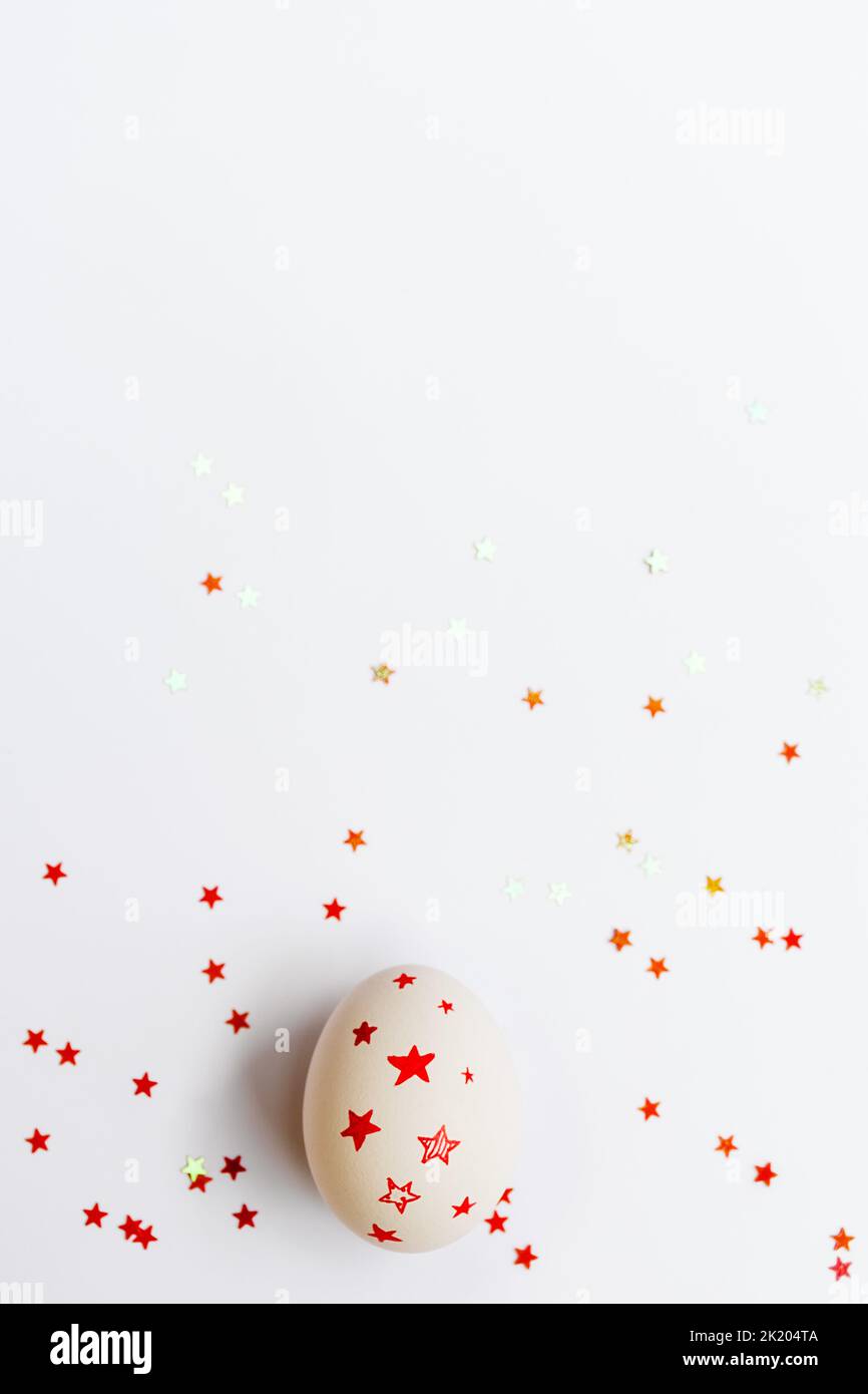 Œuf de Pâques blanc avec étoiles rouges sur fond blanc. Confettis brillants scintillants. Concept de vacances et fait main. Copier l'espace pour le texte. Photo verticale Banque D'Images