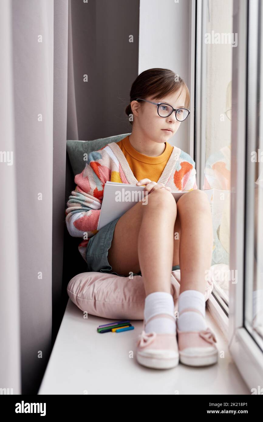 Portrait vertical de l'adolescente avec le syndrome de Down en dessinant des images tout en étant assis par la fenêtre dans la pièce confortable Banque D'Images