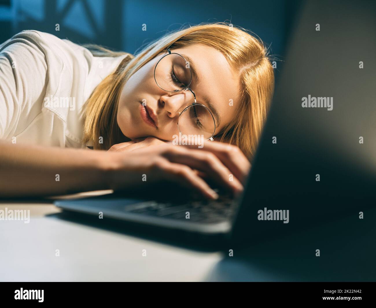 Étude de nuit. Fatigue de travail. Une étudiante en lunettes de vue calme et fatiguée dormant avec un ordinateur portable ouvert allongé sur un bureau dans un lieu de travail sombre. Banque D'Images