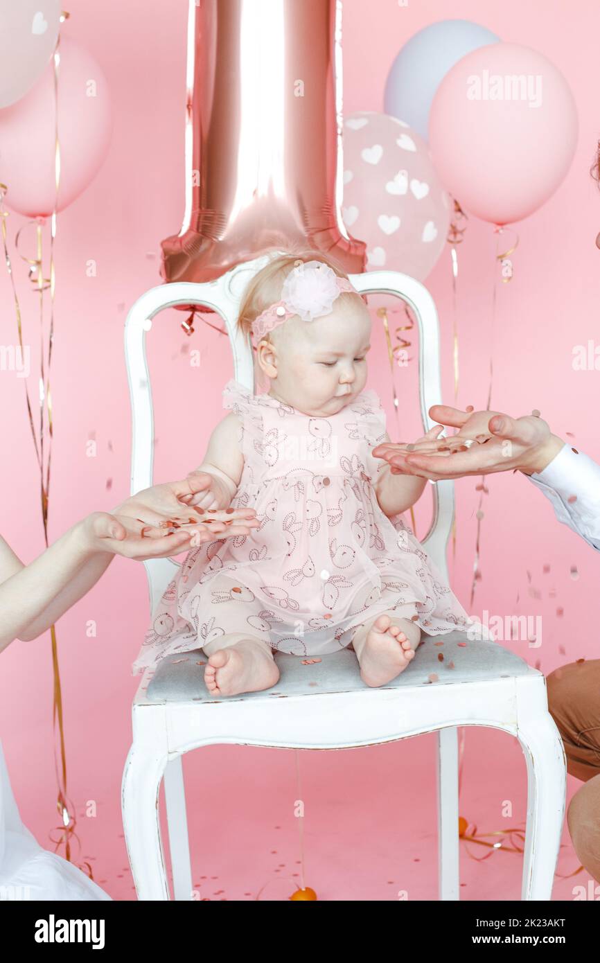 Vertical concentré un an pieds nus bébé enfant en robe rose assis sur la chaise tenir les mains des parents contre des ballons Banque D'Images