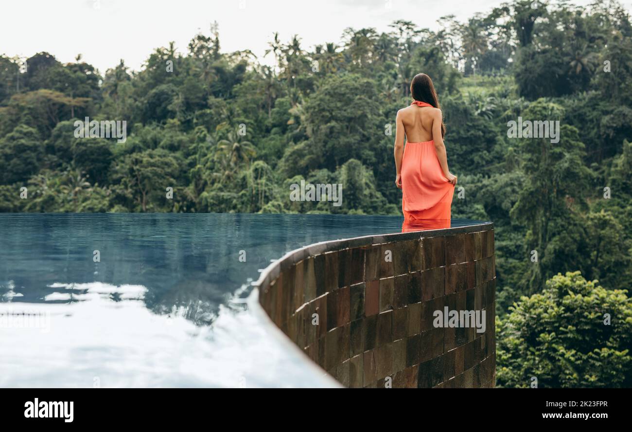 Vue arrière de la jeune femme en robe orange debout dans la piscine à débordement de la station de luxe. Banque D'Images