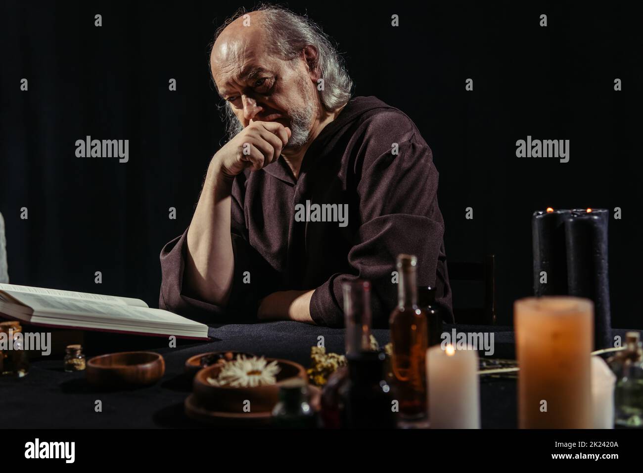 alchimiste réfléchi lisant le livre de cuisine magique près des ingrédients et des bougies isolées sur noir, image de stock Banque D'Images