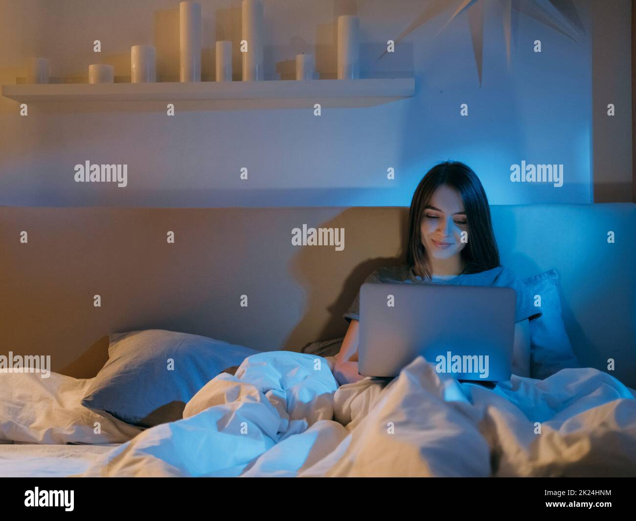 Chat vidéo de lit. Loisirs de nuit. Communication en ligne. Repos à la maison. Une femme heureuse et détendue qui s'amuse à discuter tard avec un ordinateur portable dans une chambre sombre avec une bonne nuit bleue Banque D'Images