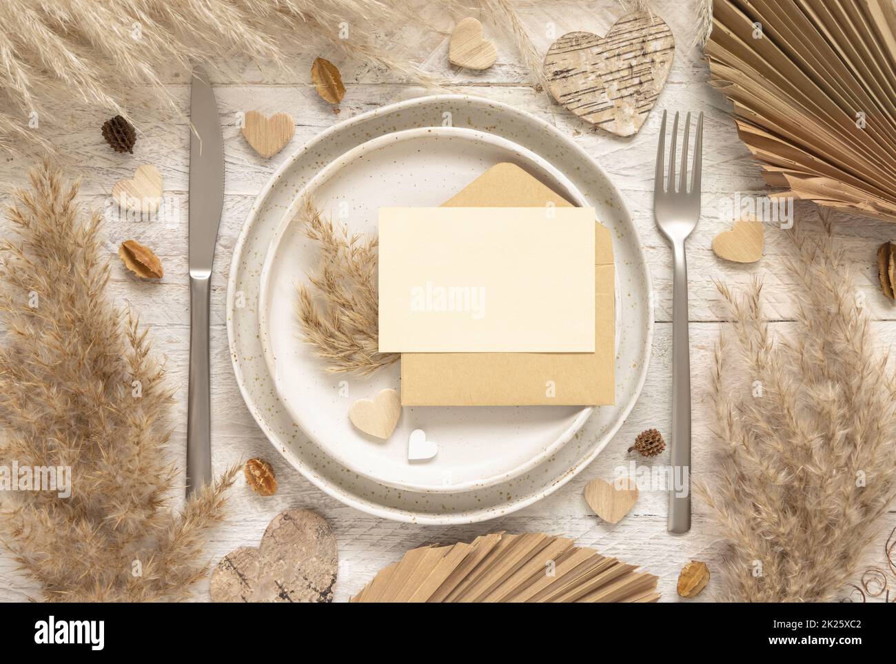 Table de mariage avec carte vierge et enveloppe près des plantes séchées et des coeurs, maquette Banque D'Images