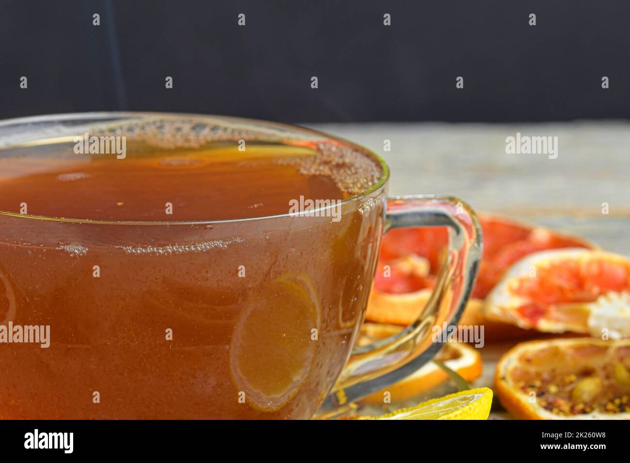 Tasse avec thé chaud et vapeur sur noir. Tasse de thé noir en verre avec bâtonnets de cannelle, étoile anisée, citron et fruits secs sur fond de table en bois blanc. Image macro Banque D'Images