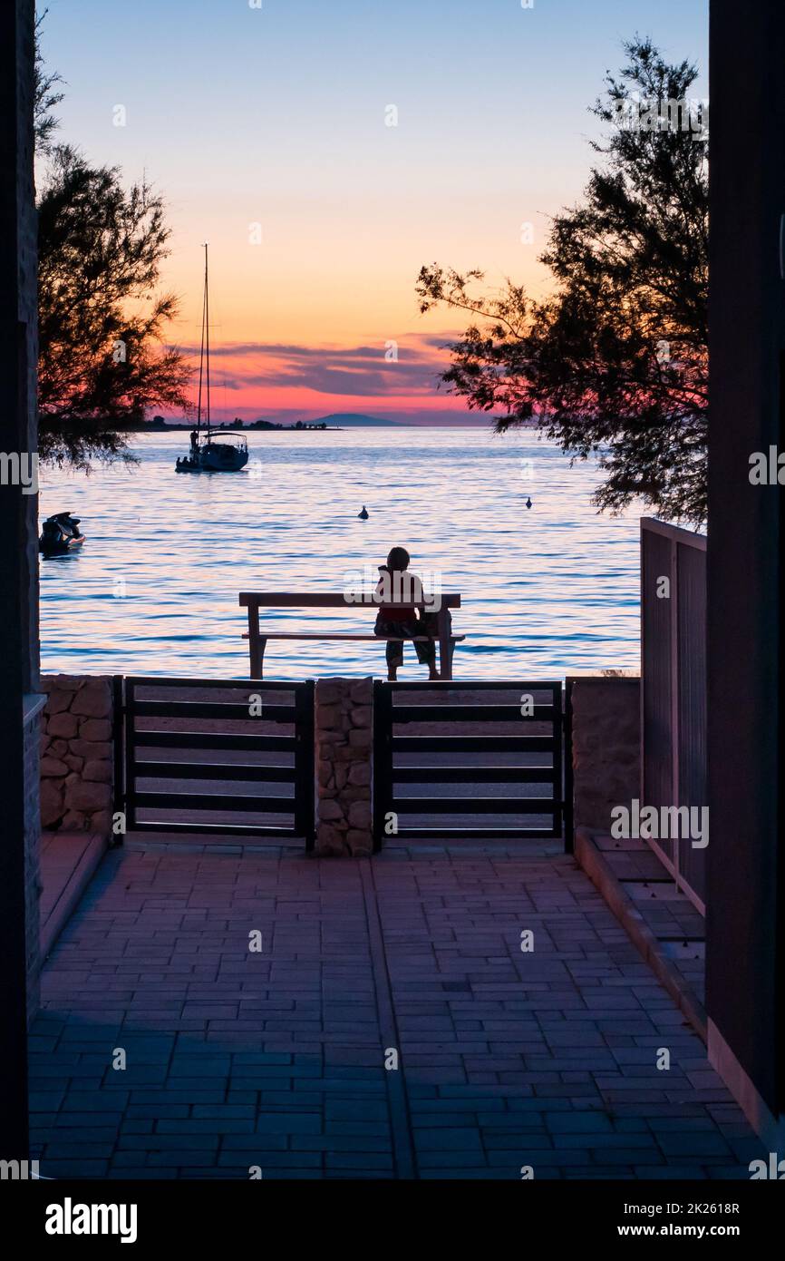 Une femme assise sur un banc près de la mer au coucher du soleil Banque D'Images