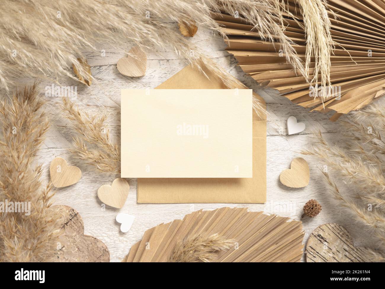 Carte vierge et enveloppe sur table en bois blanc près de l'herbe et des coeurs de pampas séchés, maquette de Boho Banque D'Images