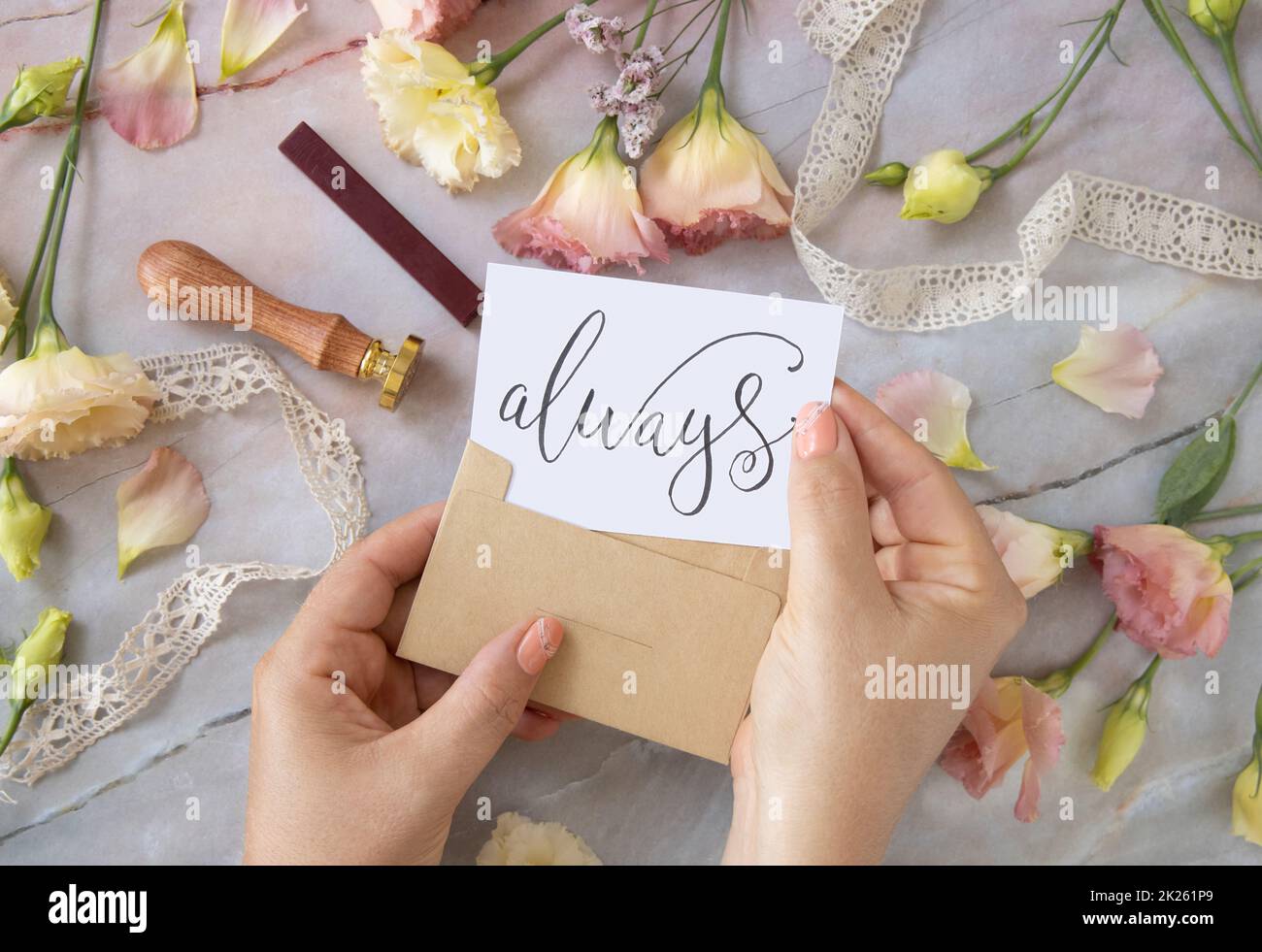Mains avec la carte TOUJOURS à l'intérieur de l'enveloppe près de fleurs roses sur une table en marbre Banque D'Images