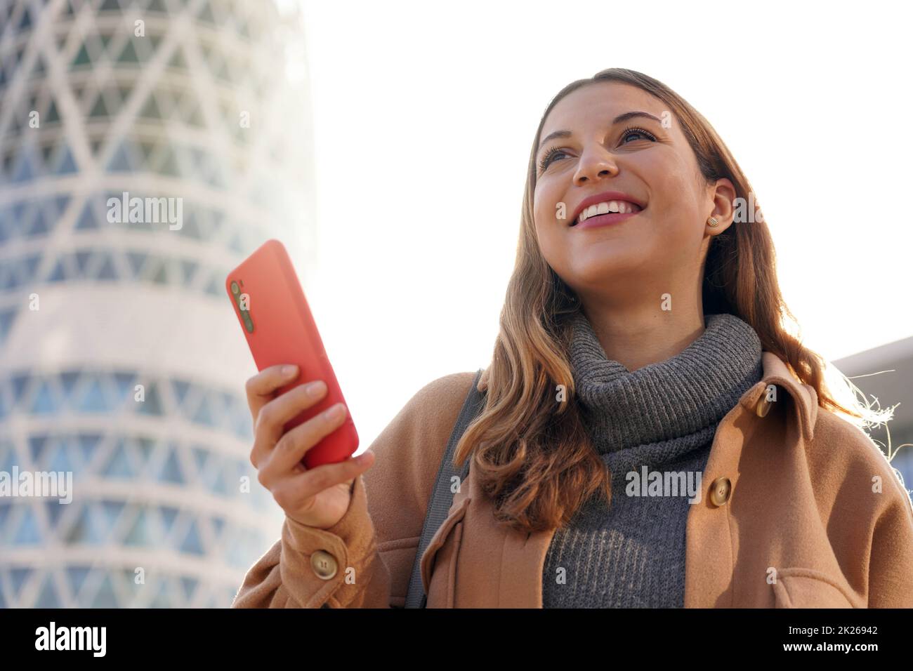 Jeune femme d'affaires confiante à la recherche d'un avenir radieux. Une belle dame excitée tenant le téléphone dans sa main avec un gratte-ciel moderne en arrière-plan. Banque D'Images