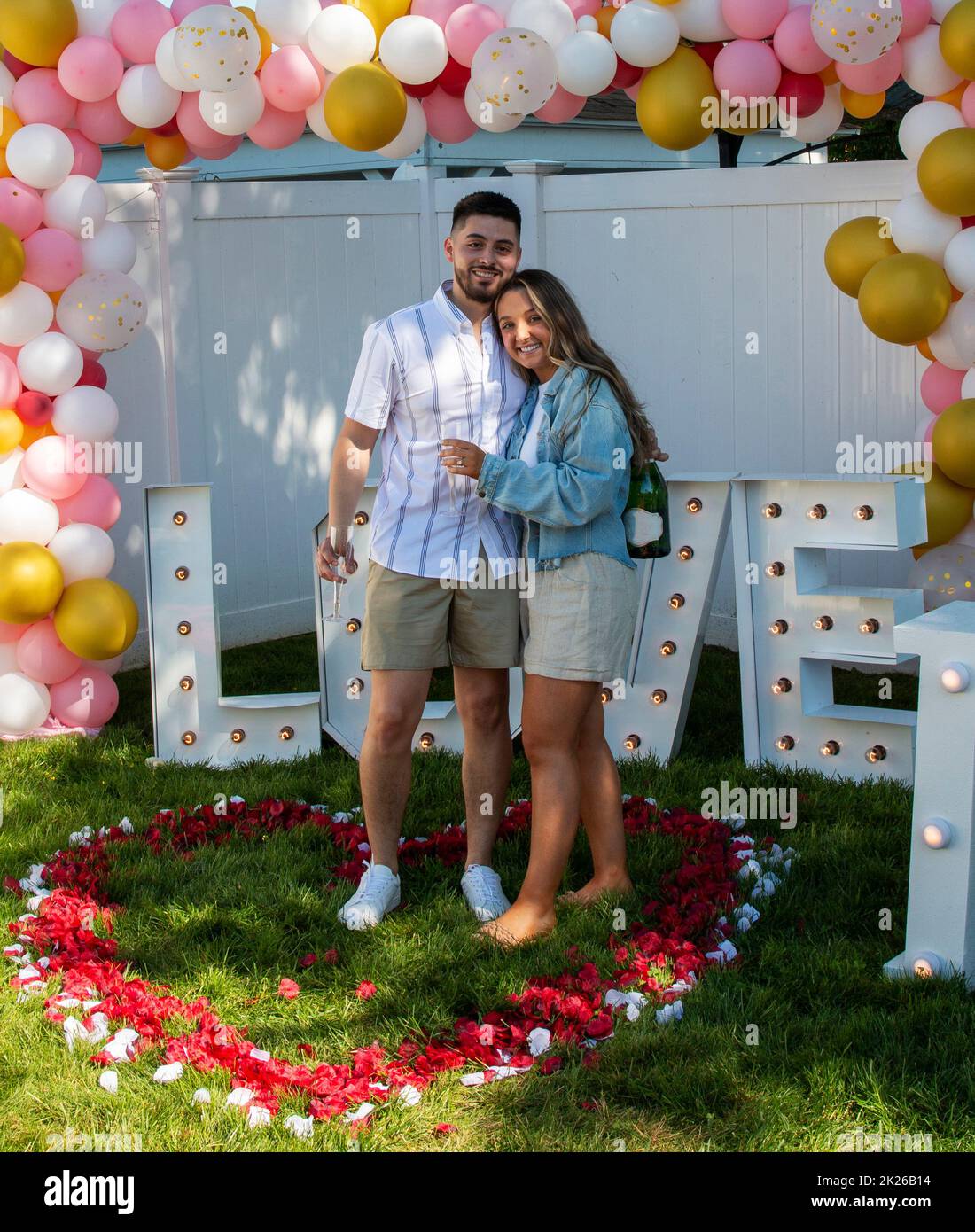Newly engaged couple standing in a heart made of red and white rose pedels with letters spelling love behind them. Banque D'Images