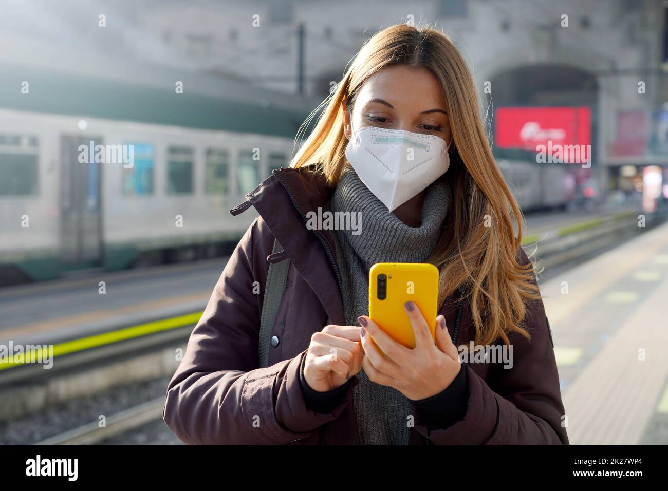 Portrait d'une fille dans un masque de protection debout sur la plate-forme de la gare avec un téléphone mobile acheter un billet en ligne Banque D'Images