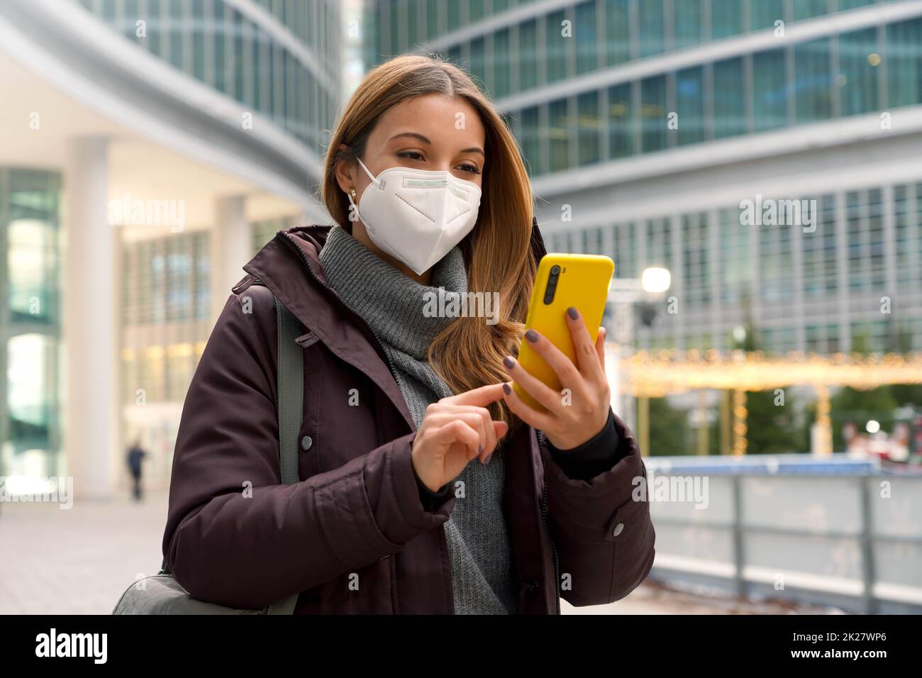 Une femme caucasienne avec des vêtements d'hiver tape avec le doigt bavardant avec quelqu'un sur le smartphone tout en portant un masque de protection à l'extérieur Banque D'Images