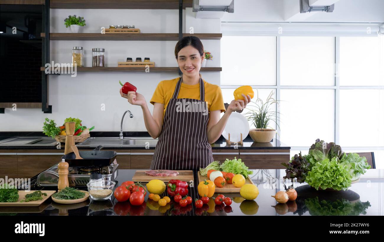 La jeune femme de ménage se tient souriante, tient le poivron rouge et jaune avec les deux mains.Le comptoir de cuisine plein de différents types de légumes. Banque D'Images