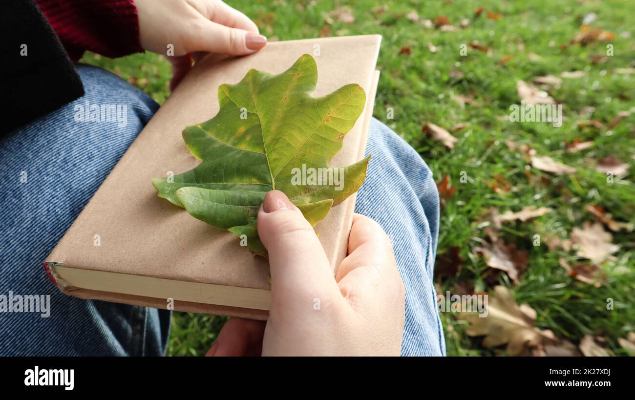 Une femme tient un livre fermé couché sur ses genoux avec une feuille de chêne tombée en gros plan dans un parc par une chaude journée d'automne ensoleillée. Le concept de détente, lecture et détente seul. Banque D'Images