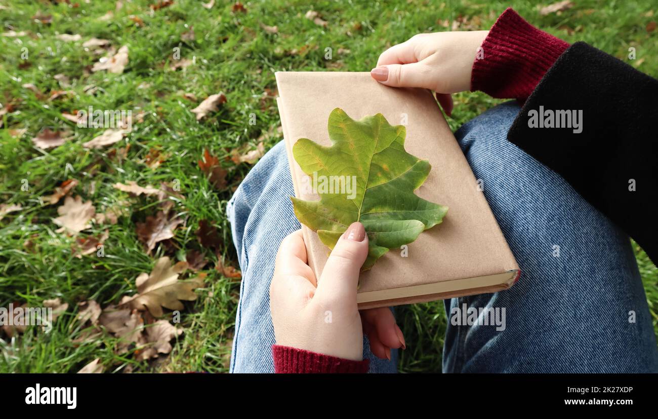 Une femme tient un livre fermé couché sur ses genoux avec une feuille de chêne tombée en gros plan dans un parc par une chaude journée d'automne ensoleillée. Le concept de détente, lecture et détente seul. Banque D'Images