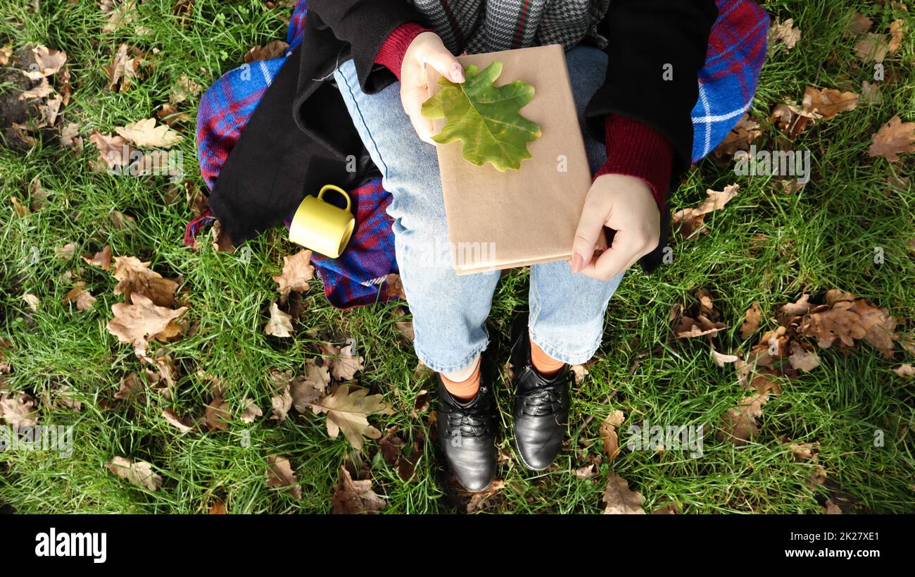 Une femme tient un livre fermé couché sur ses genoux avec une feuille de chêne tombée dans un parc lors d'une chaude journée d'automne ensoleillée sur un pré vert. Le concept de détente, lecture et détente seul. Vue de dessus, plan d'agencement. Banque D'Images