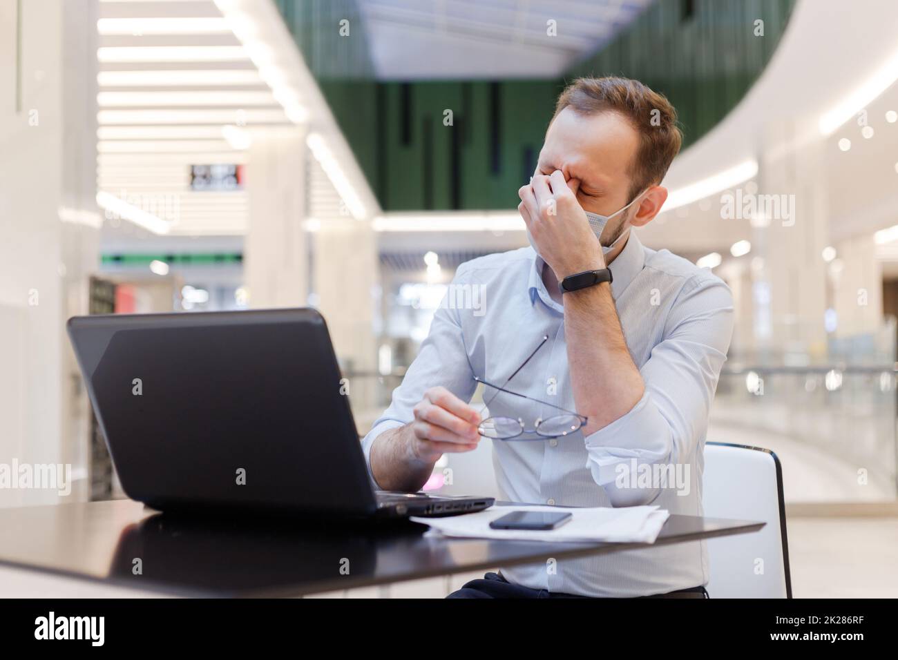 Un gars fatigué portant un masque se sent épuisé et ressent de la fatigue oculaire, touchant les yeux fermés travaillant sur un ordinateur portable Banque D'Images