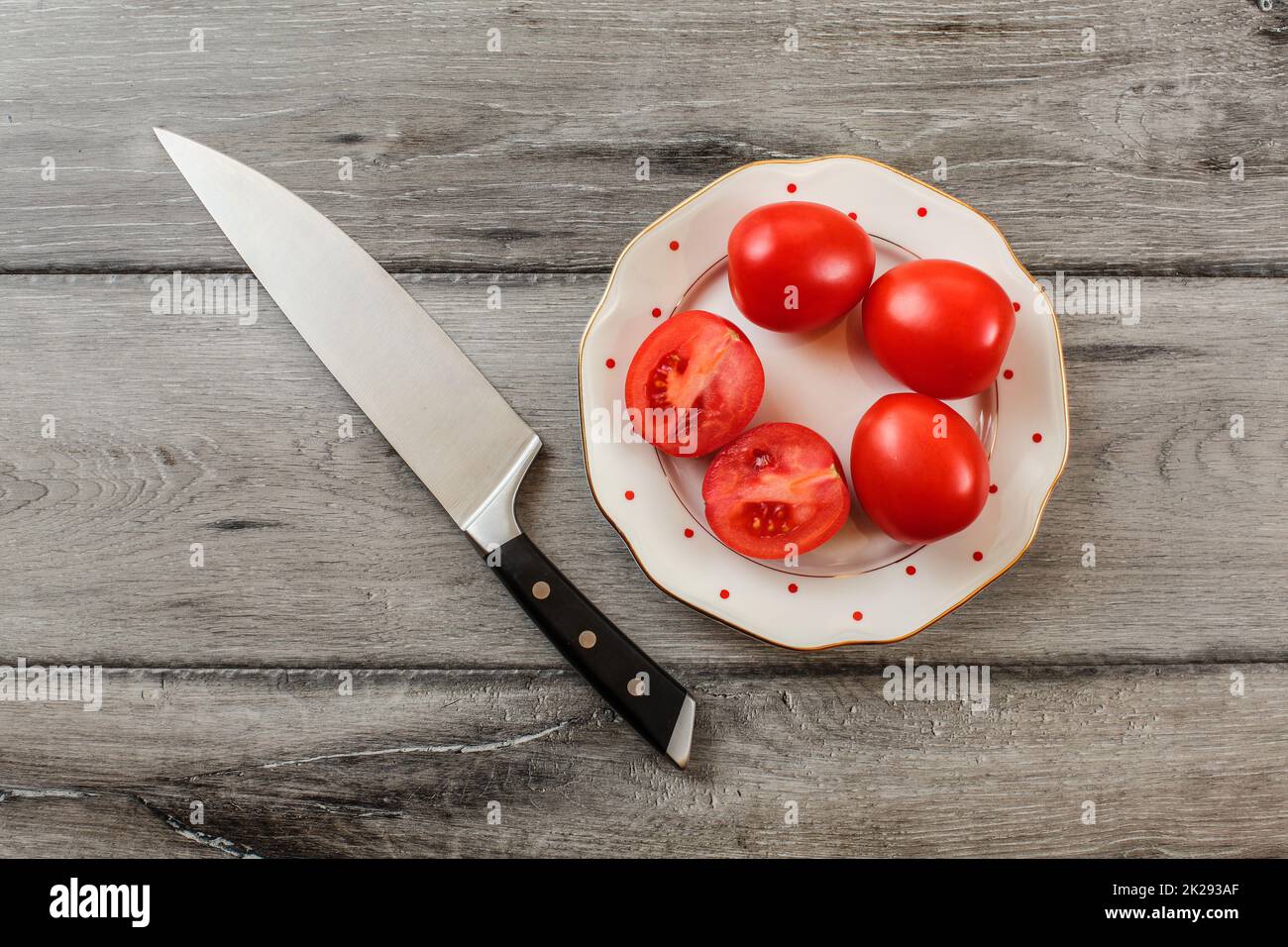 Les tomates, l'un d'entre eux réduit de moitié sur l'assiette de porcelaine blanche avec de petits points rouges, couteau chef situées près de. Vue sur table. Banque D'Images