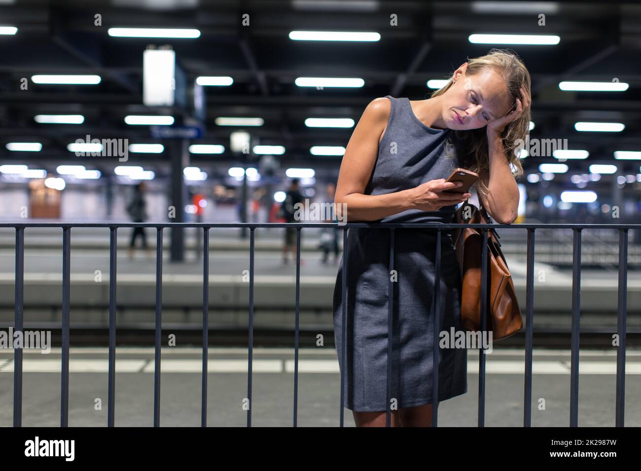 Les jeunes de banlieue surchargés épuisé femme attendant son train par jour dans une gare moderne, en utilisant son portable lors de l'attente (tons de couleur libre) Banque D'Images