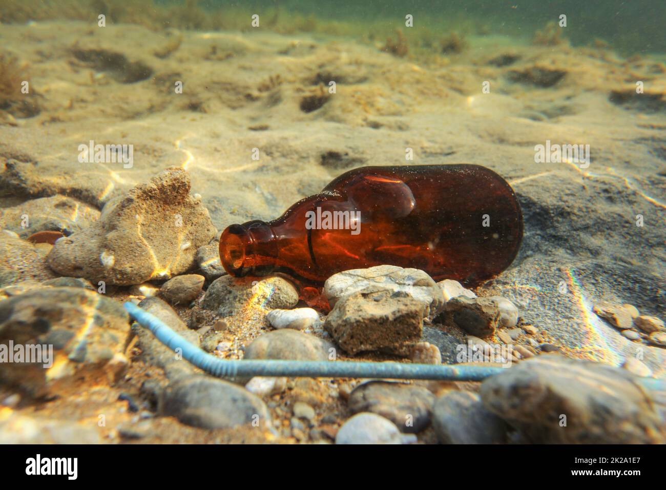 Petite bouteille de bière brune et de la paille en plastique bleu sur fond de mer de sable. Photo sous-marine, de l'océan de détritus concept. Banque D'Images