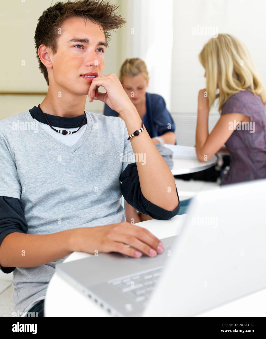 Jeune homme assis près d'un ordinateur portable. Beau jeune homme assis près d'un ordinateur portable avec des filles en arrière-plan. Banque D'Images