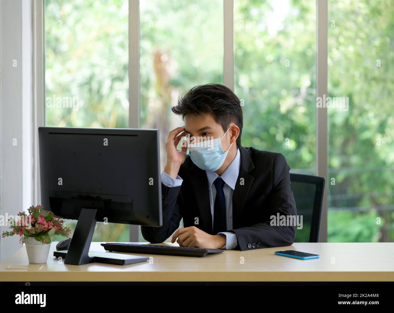De jeunes hommes d'affaires asiatiques avec masque de visage touchent son temple d'une main tout en travaillant dans le bureau. Banque D'Images