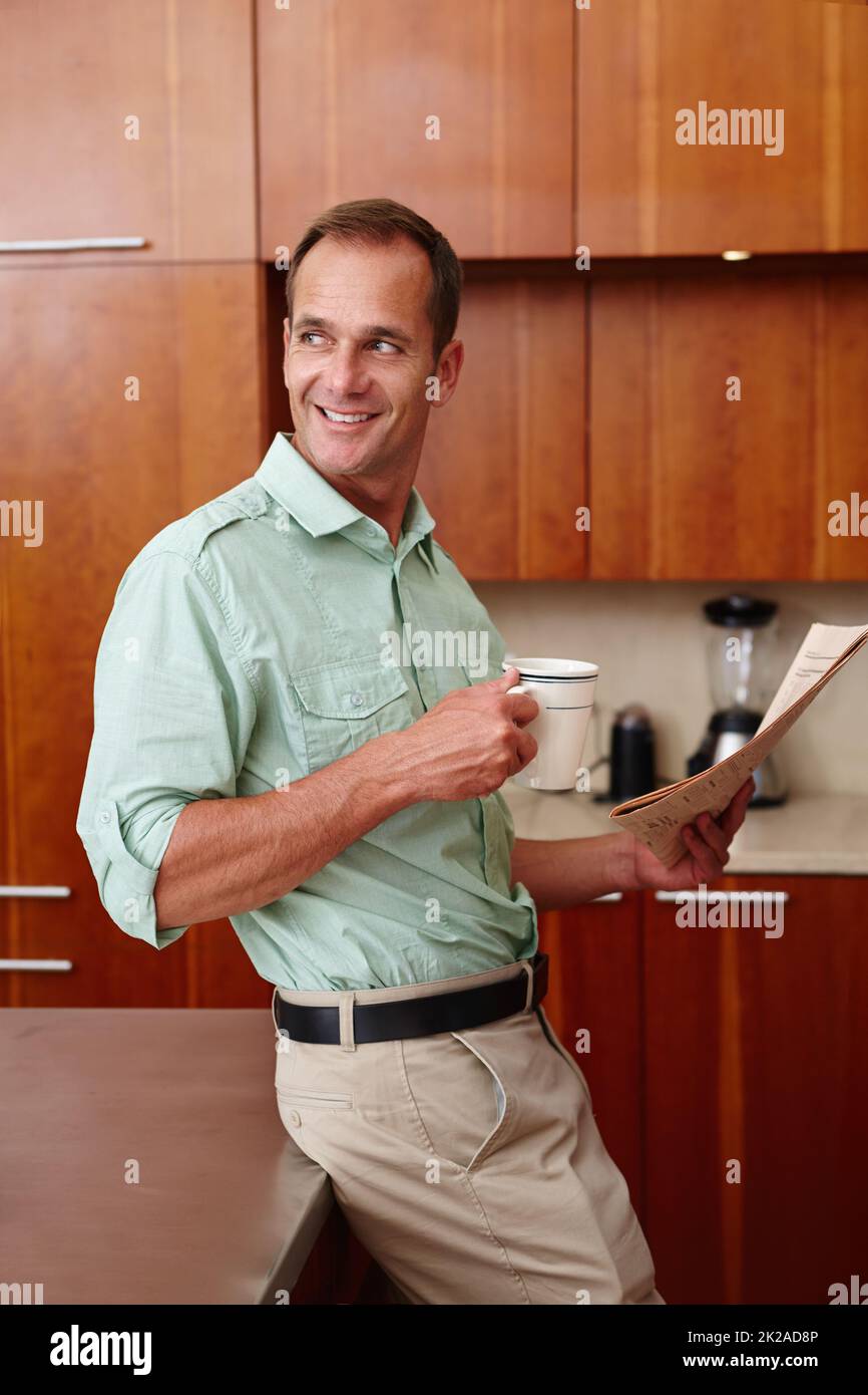 Routine du matin. Un homme heureux tenant un journal et une tasse tout en se penchant contre un comptoir dans une cuisine. Banque D'Images