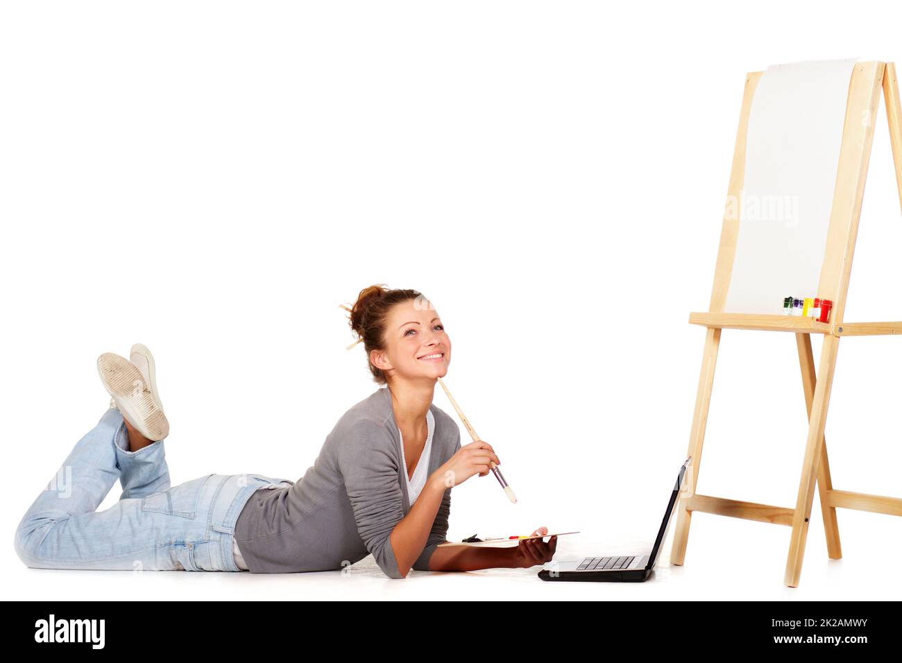 Je cherche de l'inspiration partout. Prise de vue d'une jeune femme souriante couchée par son ordinateur portable et d'une toile vierge sur un chevalet. Banque D'Images