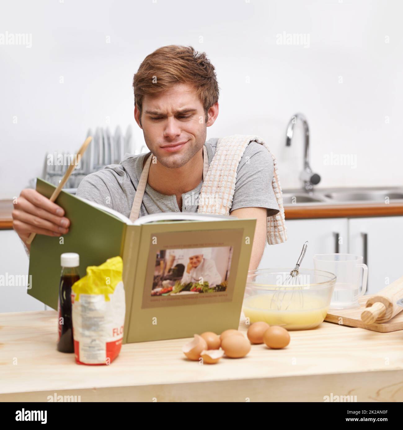 C'est un peu plus difficile que je ne l'avais prévu Un jeune homme debout dans sa cuisine et regardant perplexe en lisant un livre de recettes. Banque D'Images