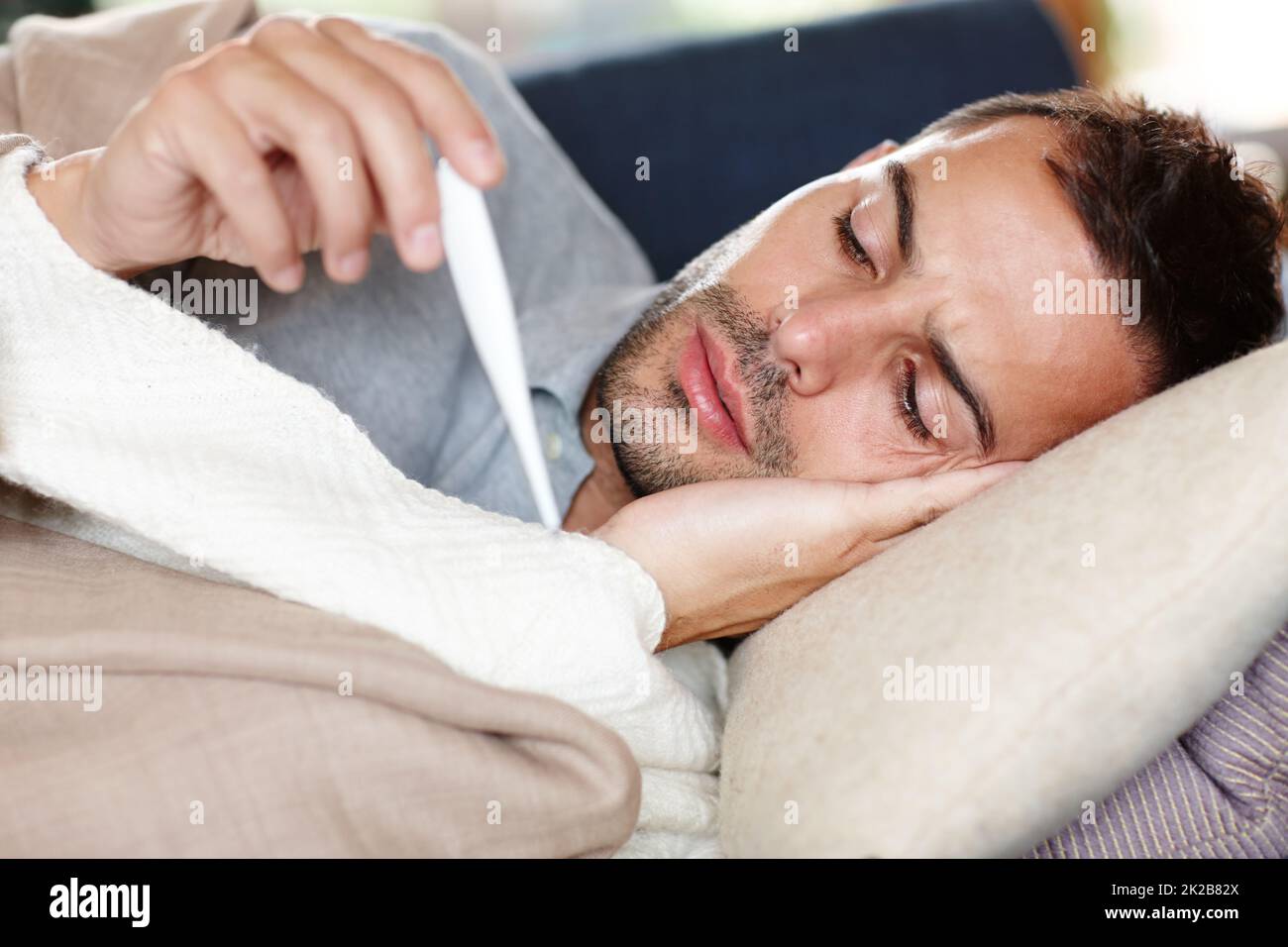 Garder un œil sur sa température. Prise de vue d'un jeune homme allongé sur le canapé et lisant sa température sur un thermomètre. Banque D'Images