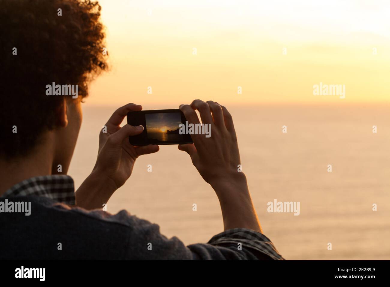 Des moments qui méritent d'être immortaliser. Vue arrière d'un jeune homme prenant une photo d'un coucher de soleil sur l'océan. Banque D'Images