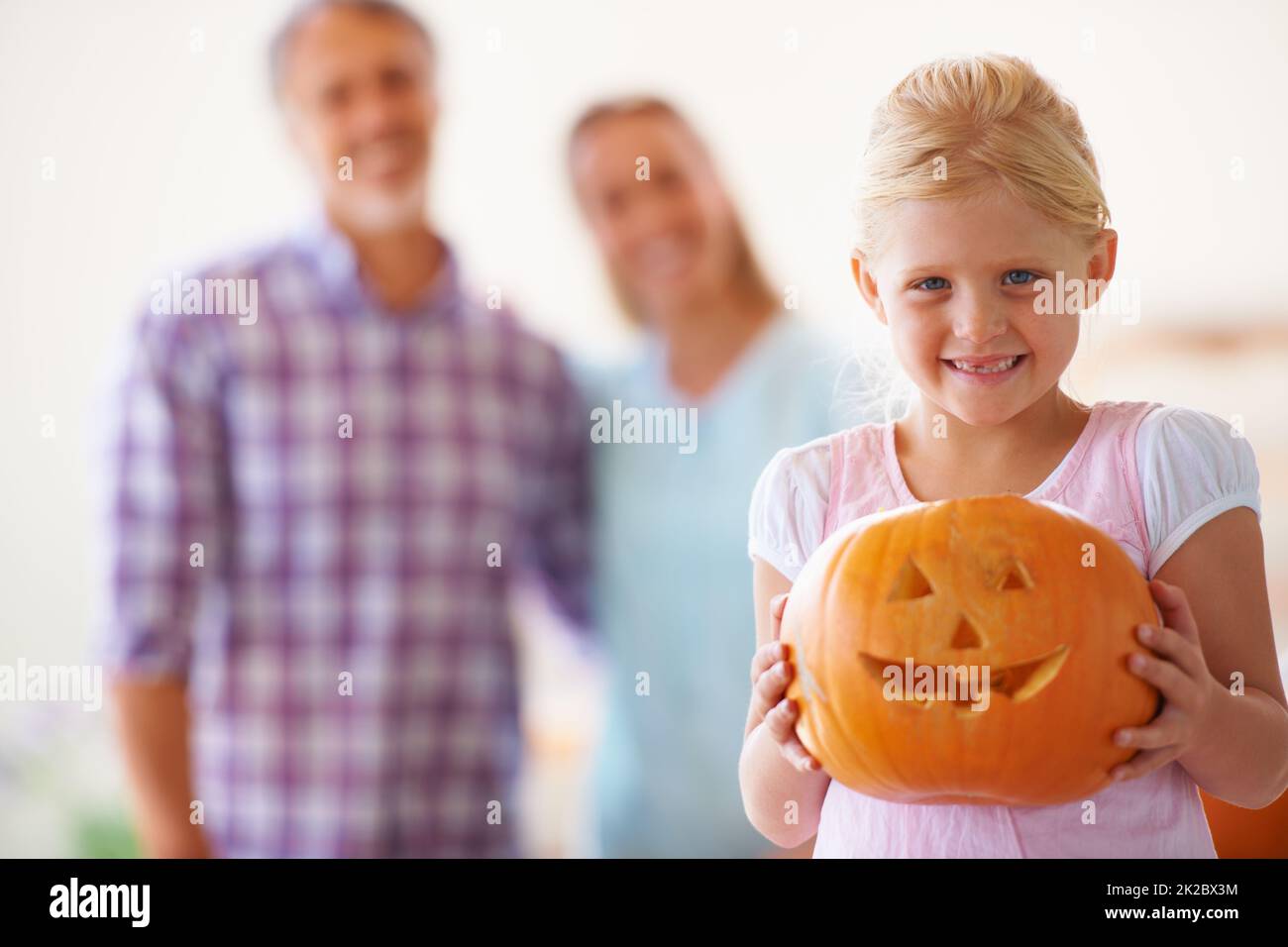 Fière de sa Jack-o-lanterne. Portrait d'une petite fille tenant sa Jack-o-lanterne avec ses parents flou derrière elle. Banque D'Images