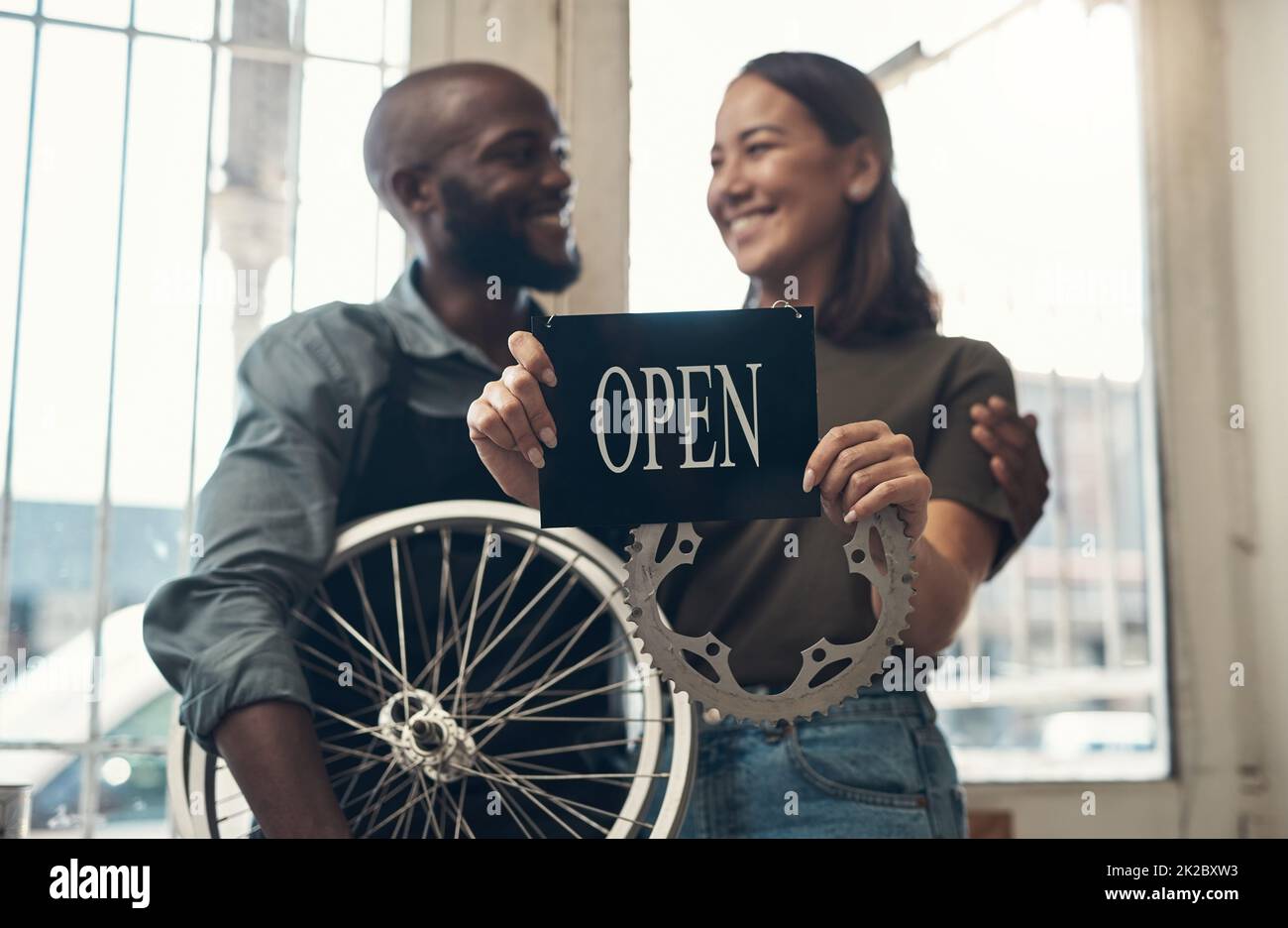 Votre vélo est entre de bonnes mains ici. Photo d'un jeune couple debout ensemble dans leur magasin de vélos et tenant un panneau ouvert. Banque D'Images