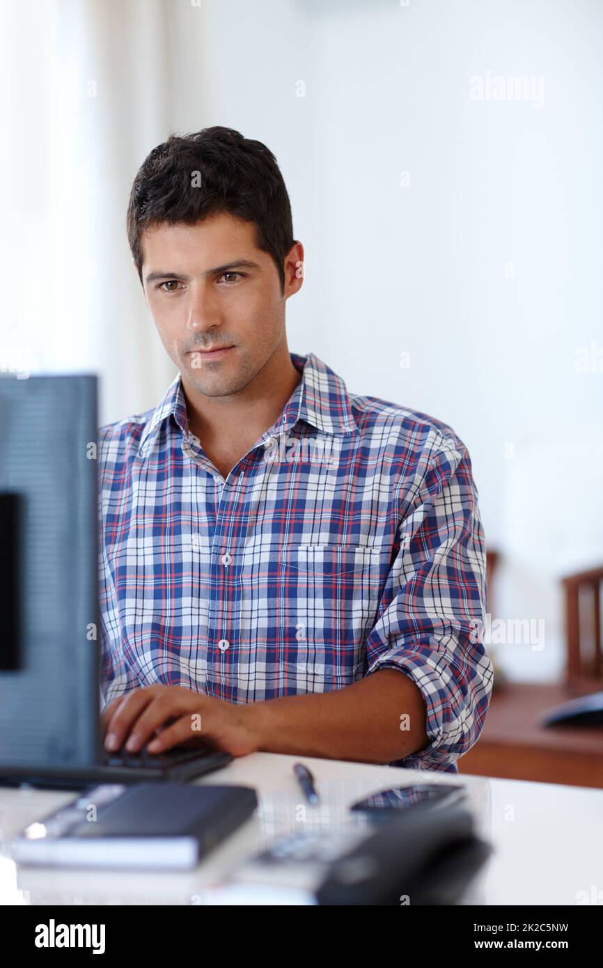 Dur au travail. Un jeune homme se concentrant tout en étant assis à un bureau devant un ordinateur. Banque D'Images
