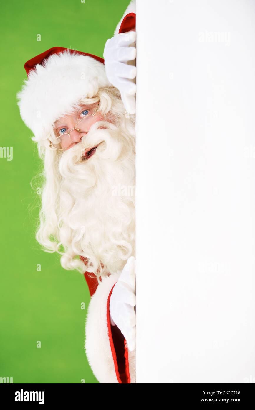 Père Noël pépirant. Santa peeking dehors autour du coin d'un mur blanc, sur un fond vert - copyspace. Banque D'Images