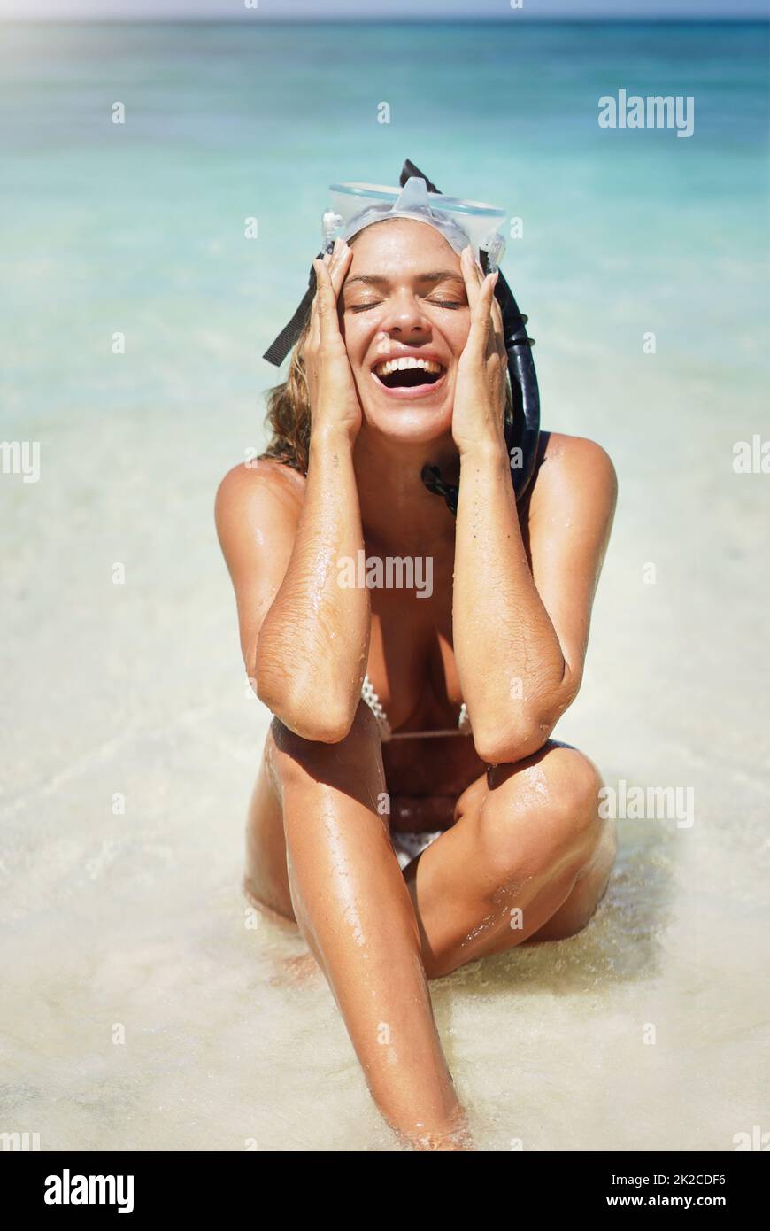 C'est toujours amusant au soleil. Photo courte d'une jeune femme attirante en équipement de plongée qui rit tout en étant assise sur la plage. Banque D'Images