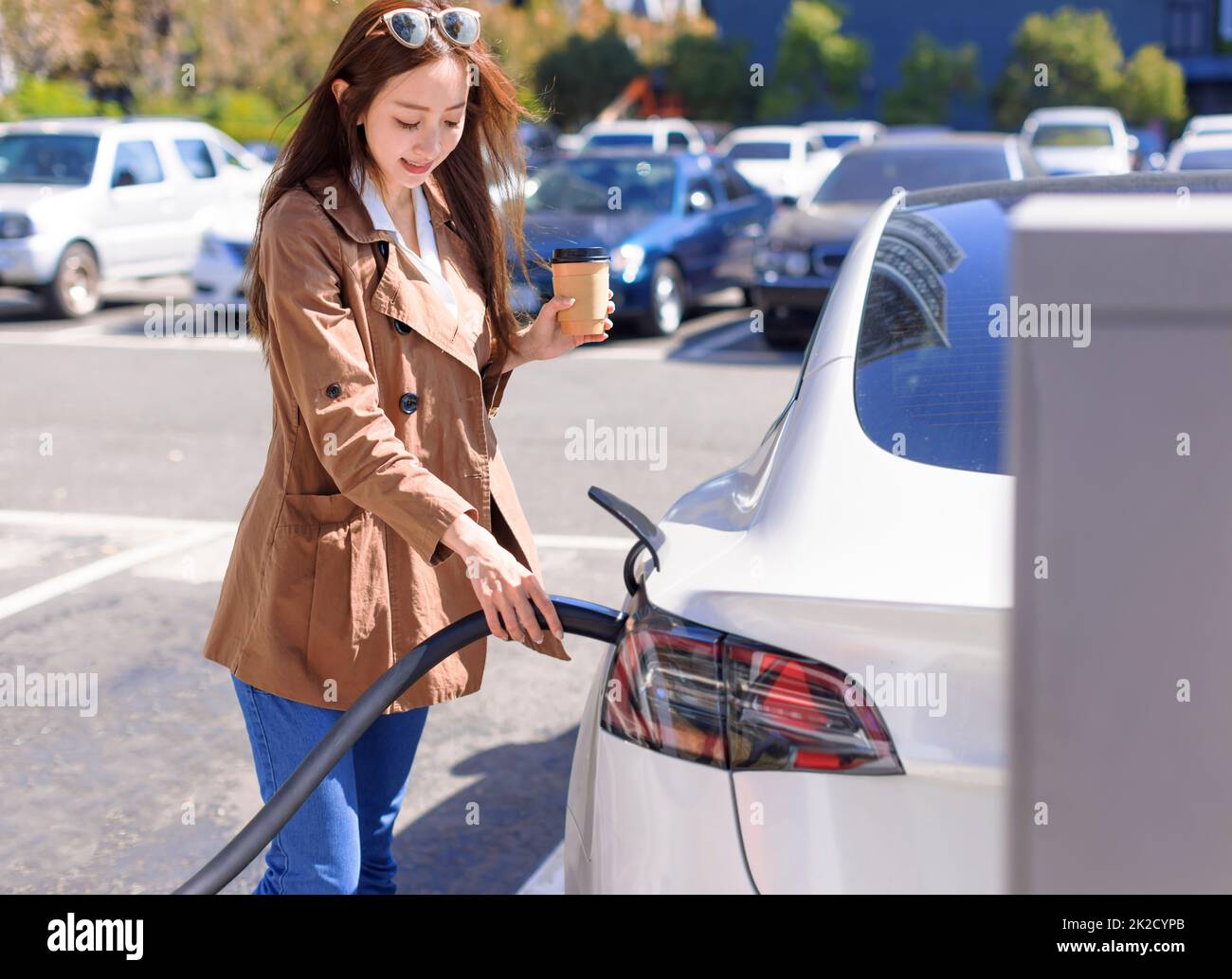 Jeune femme souriante se tenant sur le parking de la ville près de la voiture électrique, en chargeant la batterie de voiture de la petite gare de la ville Banque D'Images