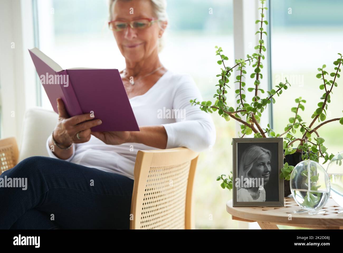 Détendu et à la retraite. Une femme âgée lisant un grand livre dans son salon, à côté d'une vieille photo d'elle-même. Banque D'Images